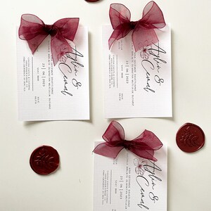 May include: Three burgundy and white wedding invitation cards with a burgundy ribbon and wax seal. The cards feature the text "Aylin & Cemal" and the date "23 | 06 | 2023".