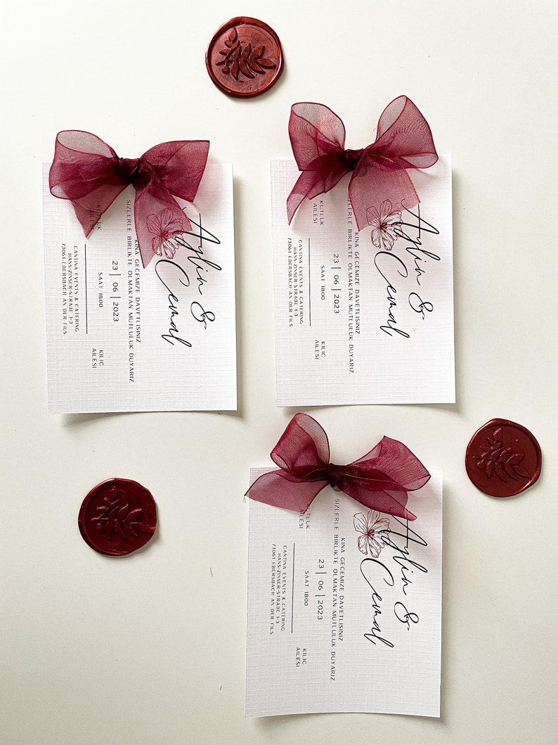 May include: Three burgundy and white wedding invitation cards with a burgundy ribbon and wax seal. The cards feature the text "Aylin & Cemal" and the date "23 | 06 | 2023".