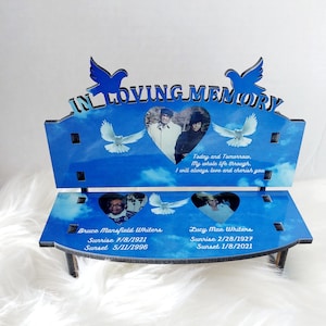 May include: A blue memorial bench with the words "IN LOVING MEMORY" and dove cutouts. It features photos in heart-shaped frames and includes names, birth, and passing dates. The bench has a sky-blue background.