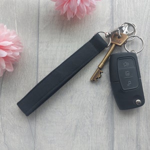 May include: A black wrist strap keyring with a silver clasp, holding a vintage brass key and a black car key fob. The keyring is made of a durable, dark fabric. Pink flowers are in the background.