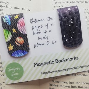 May include: Two magnetic bookmarks with a black background. One bookmark features a colourful space-themed design with planets and stars. The other bookmark has a black background with white stars. The bookmarks are displayed on a white card with the text "Between the pages of a book is a lovely place to be" and "Magnetic Bookmarks". The card also includes a website address.