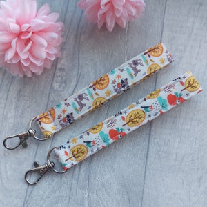 May include: Two white fabric keychains with a woodland animal print. The keychains feature a variety of animals, including raccoons, rabbits, and birds. The keychains are attached to silver key rings.