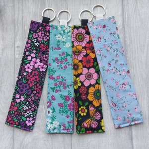 May include: Four keychains with different floral patterns. The keychains are made of fabric and have silver metal rings. The patterns include pink and black flowers on a black background, pink and green flowers on a blue background, orange and red flowers on a black background, and blue and white flowers on a blue background.