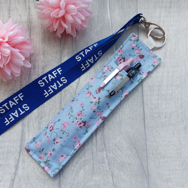May include: A blue lanyard with the word "STAFF" printed on it, attached to a light blue fabric pen holder with a floral pattern. The pen holder has two pens inside, one pink and one black.