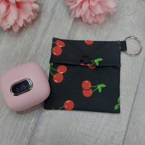 May include: A black fabric pouch with a cherry print and a snap closure. The pouch has a metal ring attached to it. A pink wireless charging case is also in the image.
