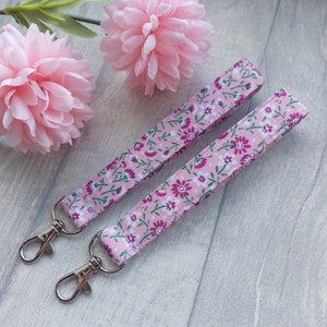 May include: Two pink floral patterned keychains with silver key rings. The keychains are made of fabric and have a pink background with white and purple flowers.
