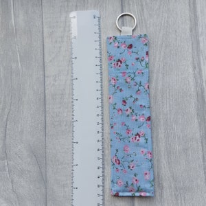 May include: A light blue fabric key chain with a floral pattern. The fabric has small pink flowers and green leaves on a light blue background. The key chain has a silver key ring.