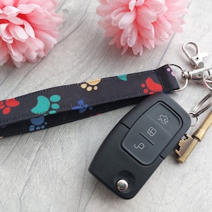 May include: A black wrist strap featuring a colourful paw print design, connected to a silver clasp and a set of keys. A black car key fob is also visible. The background includes pink floral decorations.