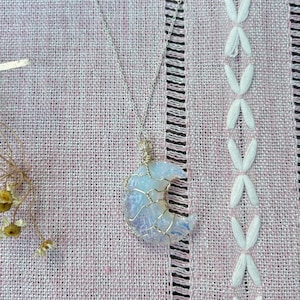 May include: A crescent moon pendant made of iridescent, semi-translucent stone wrapped in thin gold-toned wire. The pendant hangs from a delicate silver-toned chain. The moon-shaped stone displays shifting blue and white light reflections.