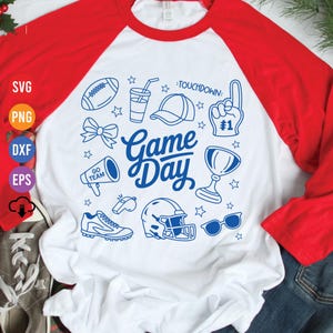 Puede incluir: Una camiseta de estilo raglán blanca y roja con un gráfico azul de "Game Day". El gráfico incluye ilustraciones relacionadas con el fútbol, como un balón, un casco y gafas de sol. La camiseta tiene mangas largas rojas y un cuerpo blanco.