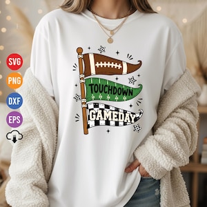 Może przedstawiać: Biały t-shirt z grafiką przedstawiającą trzy ułożone w stos proporczyki na maszcie. Górny proporczyk jest brązowy z motywem piłki do futbolu, środkowy zielony ma napis TOUCHDOWN, a dolny czarno-biały w szachownicę ma napis GAMEDAY.