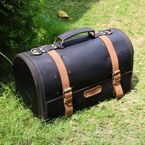 May include: Black leather travel bag with tan leather straps and handle. The bag has a rounded shape with a zipper closure and brass-colored hardware. The bag is sitting on green grass.