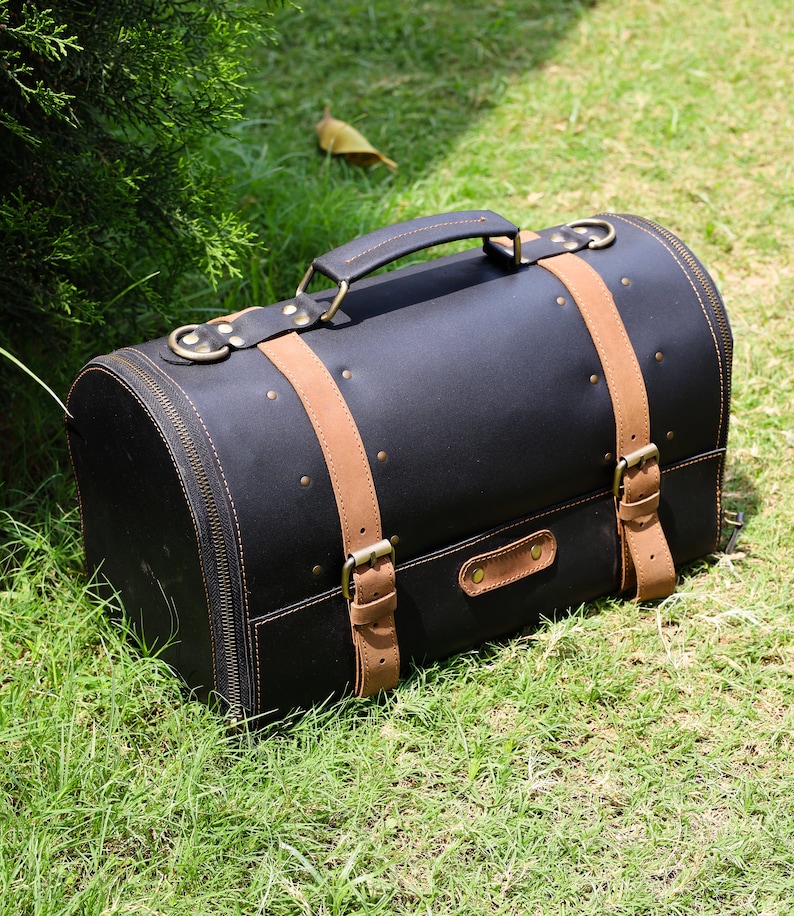 May include: Black leather travel bag with tan leather straps and handle. The bag has a rounded shape with a zipper closure and brass-colored hardware. The bag is sitting on green grass.