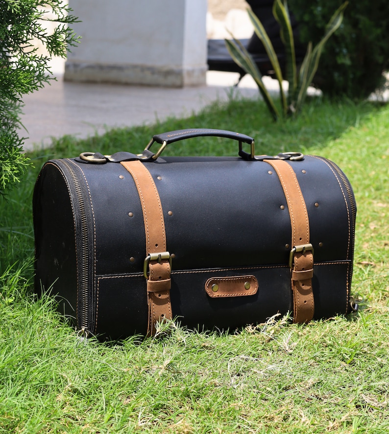 May include: Black leather satchel with tan leather straps and brass hardware. The bag has a rounded shape, a top handle, and a zipper closure. It is sitting on green grass.