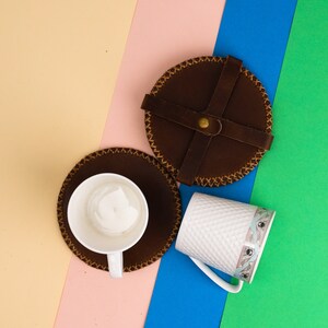 Könnte beinhalten: Zwei braune Lederuntersetzer mit genähten Kanten und eine weiße Keramiktasse mit einem dekorativen Muster. Die Untersetzer befinden sich auf einem mehrfarbigen Hintergrund.