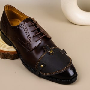 Może przedstawiać: Brązowy skórzany but oxford z czarną skórzaną końcówką i ozdobnym paskiem. But ma sznurowanie i wzór skrzydełka.