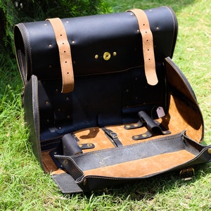 May include: Black leather satchel with tan leather straps and interior. The bag is open, revealing a tan interior with compartments and a small tray. Brass-colored rivets and hardware are visible. The satchel is sitting on green grass.