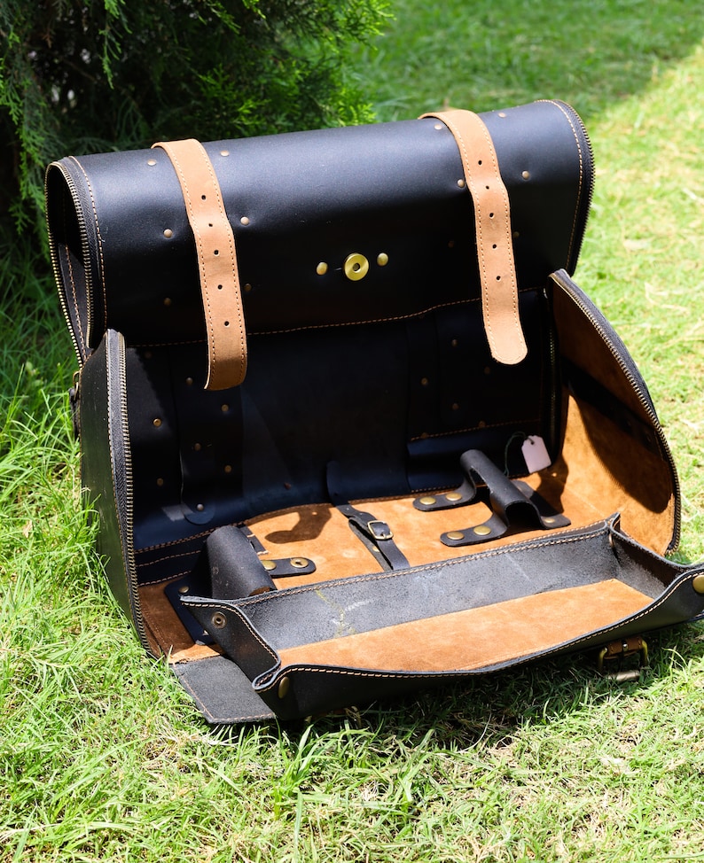 May include: Black leather satchel with tan leather straps and interior. The bag is open, revealing a tan interior with compartments and a small tray. Brass-colored rivets and hardware are visible. The satchel is sitting on green grass.