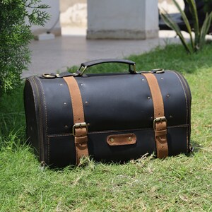 May include: A black leather satchel with tan leather straps and brass-colored hardware. The bag has a rounded shape, a top handle, and a zipper closure. It is sitting on green grass.