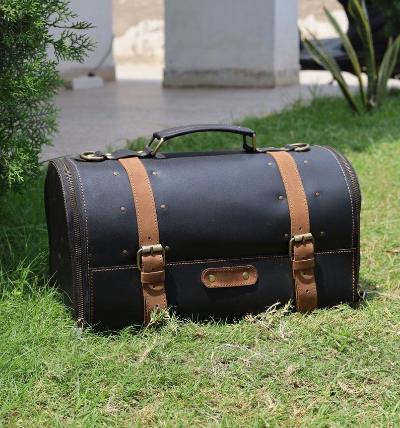 May include: A black leather satchel with tan leather straps and brass-colored hardware. The bag has a rounded shape, a top handle, and a zipper closure. It is sitting on green grass.