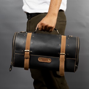May include: A black leather duffel bag with tan leather straps and gold-colored hardware. The cylindrical bag has a zipper closure and is held by a person. The bag has a classic, vintage aesthetic.