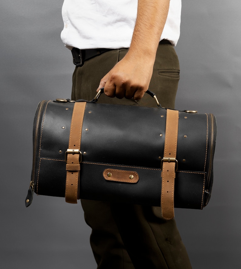 May include: A black leather duffel bag with tan leather straps and gold-colored hardware. The cylindrical bag has a zipper closure and is held by a person. The bag has a classic, vintage aesthetic.
