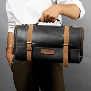 May include: Black leather duffel bag with tan leather straps and brass hardware. The cylindrical bag has a zipper closure and a top handle. The bag is held by a person.