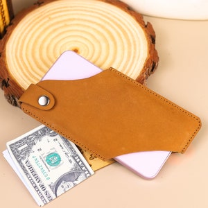 May include: A brown leather phone case with a flap and a snap closure. The case has a light pink phone inside and a dollar bill tucked into the flap.