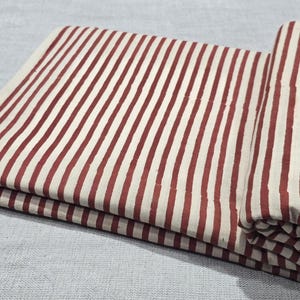 Puede incluir: Tela doblada con un patrón repetitivo de rayas verticales en color rojo intenso sobre un fondo crema. La tela está parcialmente enrollada, mostrando la continuidad del patrón. El material parece ser algodón o lino.