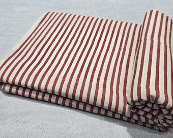 May include: Folded fabric with a repeating pattern of vertical stripes in a deep red color against a cream background. The fabric is partially rolled, showing the pattern's continuity. The material appears to be cotton or linen.