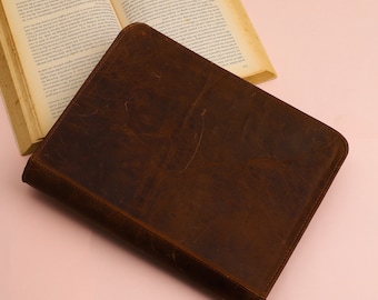 May include: A brown leather binder with a distressed finish. The binder is closed and lying flat on a pink surface.