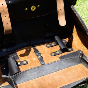 May include: Open black leather briefcase with tan leather straps and interior. The briefcase has a zippered closure and brass hardware. The interior features a tan suede-like lining and several compartments for organization.