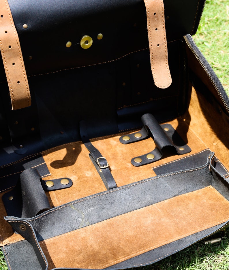 May include: Open black leather briefcase with tan leather straps and interior. The briefcase has a zippered closure and brass hardware. The interior features a tan suede-like lining and several compartments for organization.