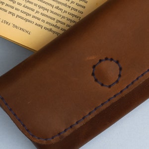 Könnte beinhalten: Eine braune Ledergeldbörse mit einem blauen, gestickten Kreisdesign. Die Geldbörse hat einen blauen, gestickten Rand.