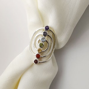 May include: Silver ring with a swirling design and seven colorful gemstones. The gemstones are arranged in a vertical line, with the largest gemstone at the top and the smallest gemstone at the bottom. The gemstones are purple, blue, green, yellow, orange, red, and brown.