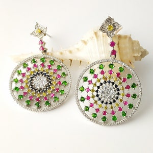 May include: A pair of silver dangle earrings with a circular design. The earrings feature a central circle of white stones surrounded by a ring of green, pink, and yellow stones. The earrings are suspended from a small flower-shaped charm.