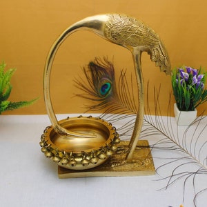 May include: A gold metal decorative stand in the shape of a crane with a bowl on top. The bowl is decorated with small bells around the rim. The stand is on a white surface with a peacock feather in the background.
