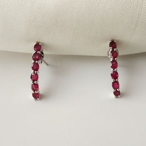 May include: A pair of silver stud earrings, each with a row of oval-cut, vivid red gemstones. The earrings are displayed against a white backdrop, highlighting the jewellery's design and colour.