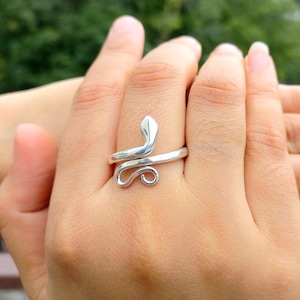Puede incluir: Un anillo plateado con forma de serpiente. El anillo se enrolla alrededor del dedo, con la cabeza de la serpiente en la parte superior y una cola enroscada en la parte inferior. El anillo se lleva en una mano, con un fondo verde borroso.