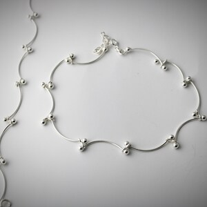 K&ouml;nnte beinhalten: Ein silbernes Fu&szlig;kettchen mit einer zarten Kette und kleinen runden Perlen. Das Fu&szlig;kettchen hat ein wellenf&ouml;rmiges Design und ist aus d&uuml;nnem Metall gefertigt.