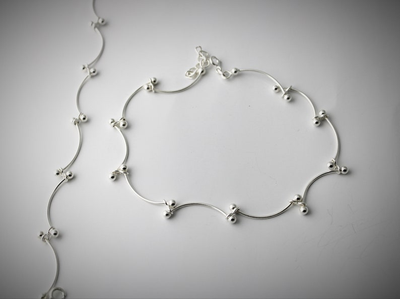 K&ouml;nnte beinhalten: Ein silbernes Fu&szlig;kettchen mit einer zarten Kette und kleinen runden Perlen. Das Fu&szlig;kettchen hat ein wellenf&ouml;rmiges Design und ist aus d&uuml;nnem Metall gefertigt.
