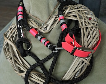 May include: Black dog leash and collar set with neon pink and silver accents. The leash is made of thick, woven black rope. The collar is neon pink with a silver buckle. The set is displayed on a woven heart-shaped decoration.