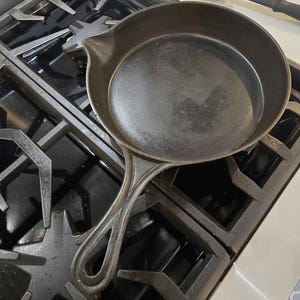 May include: A round, black cast iron skillet with a smooth interior surface and a long, hollow handle featuring a teardrop-shaped opening. The pan has a small pouring spout on the side and a textured exterior finish, resting on a stove grate.