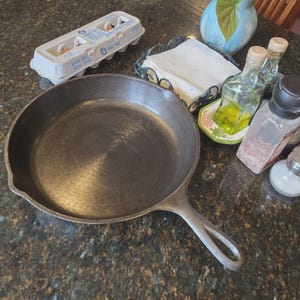May include: A well-used cast iron skillet with a long handle sits on a countertop. Nearby are a carton of eggs, a napkin holder, oil and vinegar bottles, and salt and pepper shakers. The skillet is dark gray.