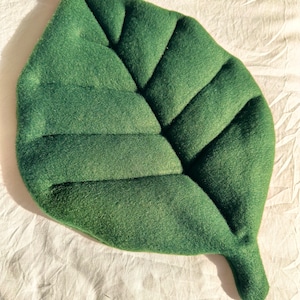 Pode incluir: Uma almofada verde em forma de folha feita de tecido macio. A almofada tem um padrão de veias detalhado e é colocada sobre uma superfície branca texturizada. A almofada tem aproximadamente 46 cm de comprimento e 36 cm de largura.