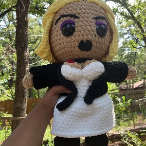 May include: Crocheted doll with blonde hair, purple eyeshadow, and black lips. The doll wears a black jacket over a white dress. The doll's eyes are large and black. The doll is handmade and has a unique, whimsical design.