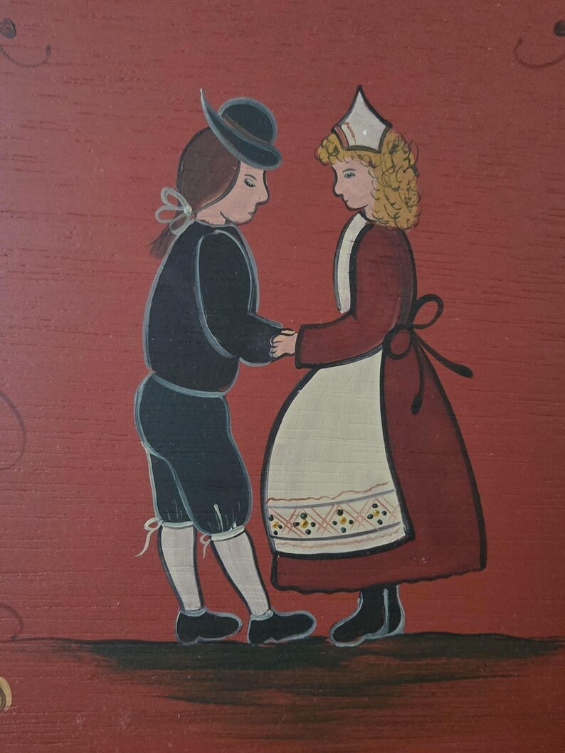 May include: A folk art painting on a red background depicts a couple in traditional attire holding hands. The man wears a black hat, jacket, and shorts. The woman is in a red dress with a white apron. The artwork has a simple, charming style.