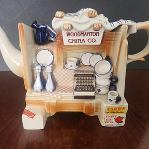 Könnte beinhalten: Eine Keramik-Teekanne in Form einer Ladenfront mit einem Schild, auf dem "Woodmanton China Co." steht. Die Teekanne ist mit verschiedenen Details verziert, darunter eine Katze, eine Teetasse und eine Teekanne.