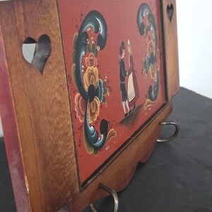 May include: Wooden wall hanging with a red background featuring a painted scene of two figures in traditional attire. The frame is brown with heart-shaped cutouts and decorative floral elements. Metal ring hardware is attached to the bottom.