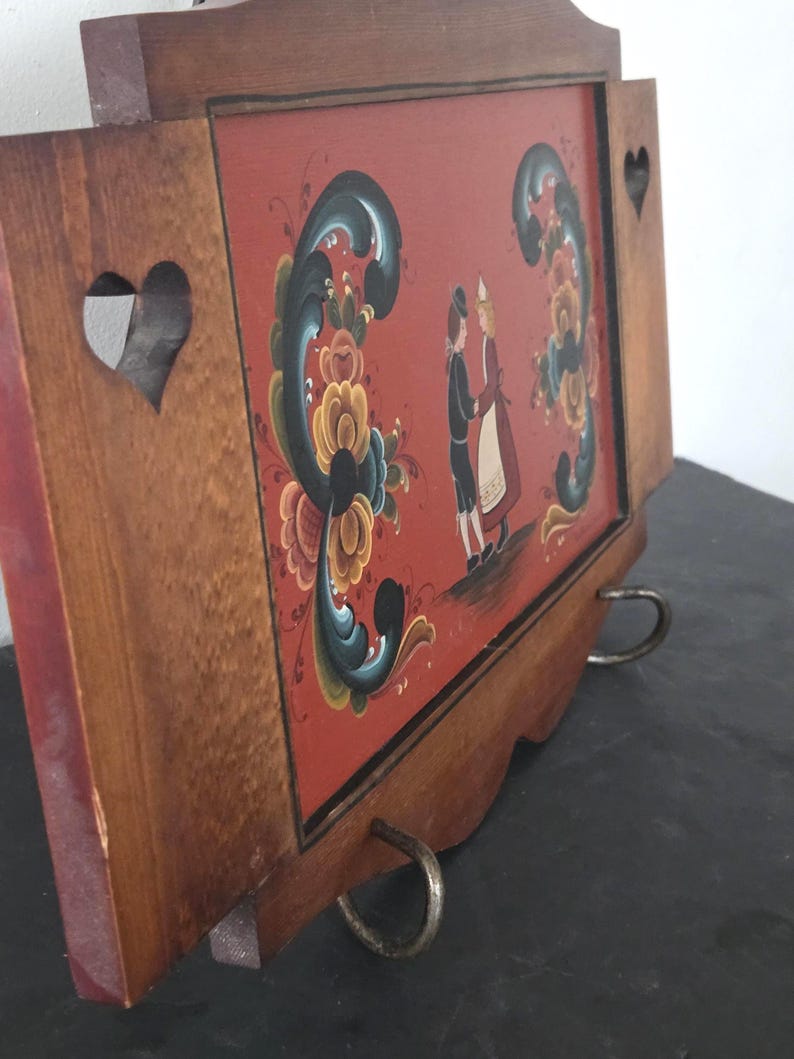 May include: Wooden wall hanging with a red background featuring a painted scene of two figures in traditional attire. The frame is brown with heart-shaped cutouts and decorative floral elements. Metal ring hardware is attached to the bottom.